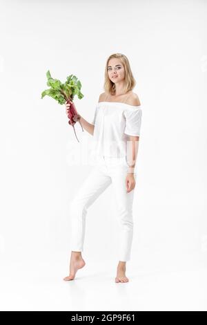 Schöne blonde Frau in einer weißen Bluse hält eine rote Bete mit grünen Blättern auf weißem Hintergrund. Gesundheit und Vitamine Stockfoto