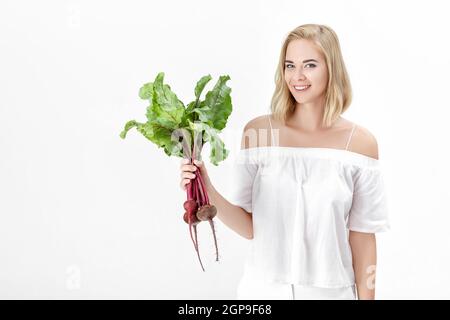 Schöne blonde Frau in einer weißen Bluse hält eine rote Bete mit grünen Blättern auf weißem Hintergrund. Gesundheit und Vitamine Stockfoto