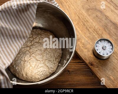 Frischer Teig in einer Metallschüssel für ein hausgemachtes Baguette auf einer hölzernen Küchentheke mit Wecker. Top-View-Fotografie. Stockfoto