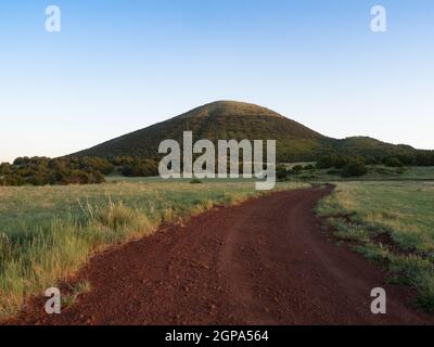 Ein Blick in den unteren Winkel auf eine rote Schotterstraße, die zum Capulin Volcano National Monument führt, mit Präriegras auf beiden Seiten und wolkenlosem Himmel darüber. Stockfoto