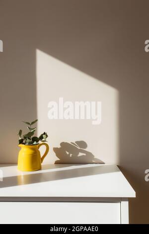 Ein gelber Keramikkrug oder eine Vase mit Eukalyptuszweigen, leere weiße Bilderrahmen auf dem weißen Tisch im Innenraum mit beigen Wänden in der Nähe des Fensters. Stockfoto