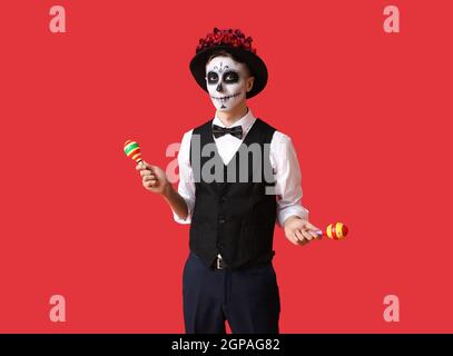 Junger Mann mit bemaltem Schädel auf dem Gesicht und Maracas vor farbigem Hintergrund. Feier des mexikanischen Tages der Toten (El Dia de Muertos) Stockfoto