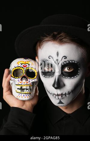 Junger Mann mit gemaltem Gesicht und Zuckerschädel für Mexikos Tag der Toten (El Dia de Muertos) auf dunklem Hintergrund Stockfoto