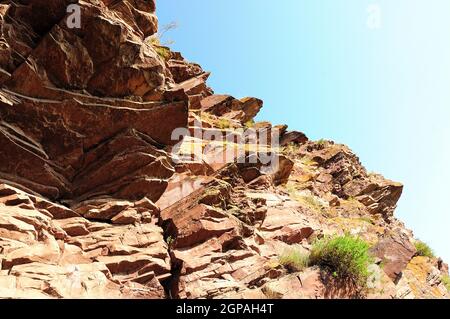 Eine schichtweise Gebirgsformation von leuchtend roter Farbe mit kleinen Büschen auf einem Hügel mit Blick auf den klaren Sommerhimmel. Bergkisten, Chakassien Stockfoto