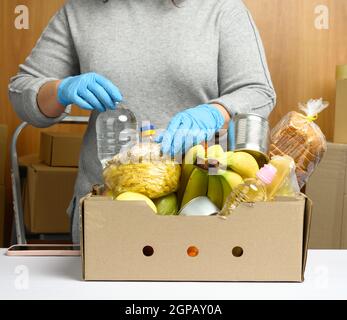 Frau in Handschuhen sammelt immer wieder Lebensmittel, Früchte und Dinge und einen Karton für die Hilfe für Bedürftige, das Konzept der Hilfe und Freiwilligenarbeit. Lieferung Stockfoto