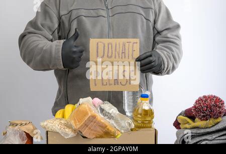 Mann hält ein Blatt Papier mit einem Schriftzug spenden bitte und sammelt Lebensmittel, Früchte und Dinge in einem Karton, um die Bedürftigen und die Armen, die zu helfen Stockfoto