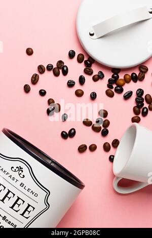 Kaffeebohnen aus der Kaffeekanne mit dem Deckel daneben und einer weißen Tasse auf einem rosa Hintergrund. Stockfoto