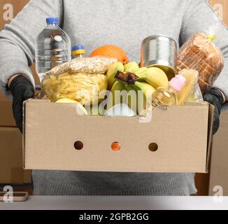 Frau in grauem Pullover und schwarzen Handschuhen mit einem Karton mit Lebensmitteln, Konzept der Unterstützung und Freiwilligenarbeit, Lieferung von Lebensmitteln Stockfoto