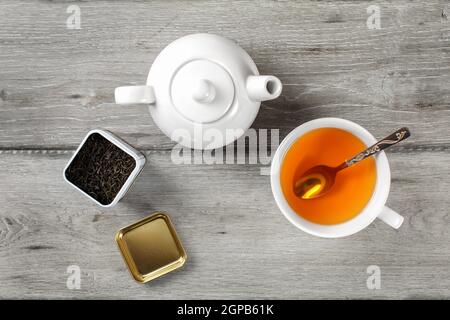 Table Top - Messing Tee Caddy, weißes Porzellan Teekanne und Tasse aus Keramik mit silbernen Löffel und heiße Amber trinken, auf grau Holzschreibtisch. Stockfoto