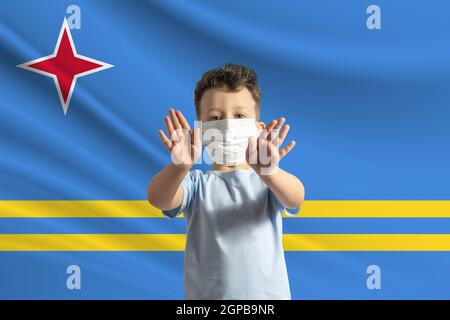 Kleiner weißer Junge in einer Schutzmaske auf dem Hintergrund der Flagge von Aruba. Macht ein Stoppschild mit seinen Händen, zu Hause bleiben Aruba. Stockfoto