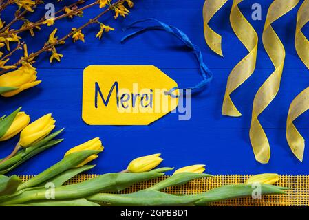 Etikett mit französischem Text Merci bedeutet Danke. Gelb Frühling Blumen Wie Tulpe Und Zweige. Festliche Dekoration Mit Band. Blauer Holzhintergrund Stockfoto
