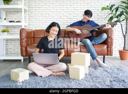 Junge Liebhaber verbringen Zeit zusammen auf Urlaub im Wohnzimmer. Asiatischer Mann mit Kopfhörern spielt eine akustische Gitarre, während das Mädchen das Paket überprüfen Stockfoto