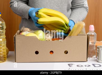 Frau in Handschuhen sammelt immer wieder Lebensmittel, Früchte und Dinge und einen Karton für die Hilfe für Bedürftige, das Konzept der Hilfe und Freiwilligenarbeit Stockfoto