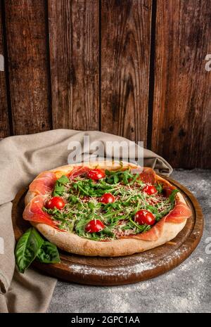 Pizza mit Prosciutto arugula und Tomaten auf Holzbrett. Pizza auf einem grauen Tisch auf einem Hintergrund aus Holzbrettern und grauem Stoff. Stockfoto