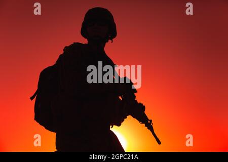 Silhouette des Armeekrieges in Helm und Munitionsform, mit taktischem Rucksack, mit Dienstgewehr auf dem Hintergrund des Sonnenuntergangs. Zählerstand Stockfoto