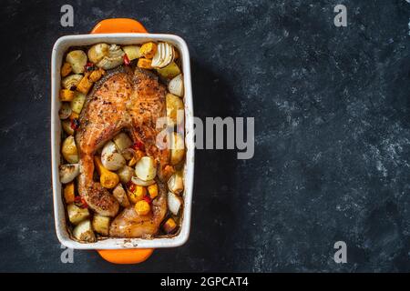 Gebackenes Lachsfilet mit Kartoffeln, Karotten, Zwiebeln, roten Paprika und Knoblauch in einer Keramikschale auf schwarzem Hintergrund, Platz kopieren, Nahaufnahme Stockfoto