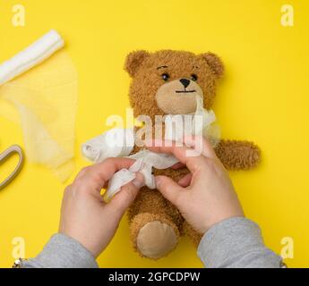 Lustiger Vintage brauner lockiger Teddybär mit aufgewickelter Pfote mit weißer Gaze-Bandage auf gelbem Hintergrund, Konzept von Verletzungen bei Kindern oder Tieren, Polizist Stockfoto