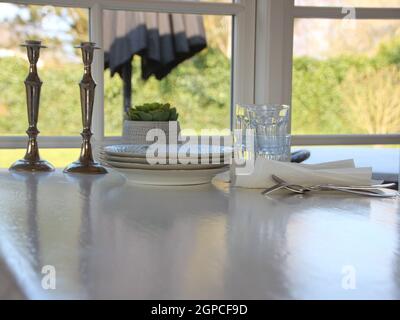 Ein alter weißer Tisch, der kurz vor dem Set steht, mit Tellern, Gläsern und Besteck. Im Hintergrund ist ein Rasen zu sehen. Stockfoto