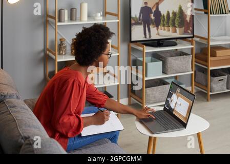 Junge Frau, die mit einem Laptop auf der Couch sitzt, Online-Mathenkurs macht und sich Notizen macht Stockfoto