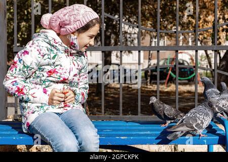 Teenager Mädchen Fütterung Tauben im Park. Das Mädchen sitzt auf einer Bank und füttert die Vögel mit Samen. Herbstsonniger Tag. Die Pflege der Vögel in einer Stadt Stockfoto