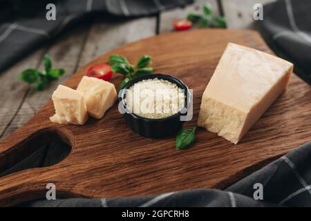 Italienisches Küchenkonzept. Schneidebrett, Parmesankäse, geriebener Käse in kleiner schwarzer Schüssel, Kirschtomatos, Basilikumblätter Zweige, Küchentücher Stockfoto