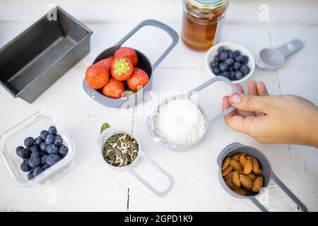 Erdbeeren, Heidelbeeren, Mehl, Mandeln und Samen in Plastikbehältern. Weibliche Hand hält eine Messbecher Mehl über dem weißen Zähler. Gesund mich Stockfoto