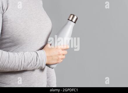 Frau in grauem T-Shirt mit grauer Isolierflasche auf grau Nahaufnahme des Hintergrunds Stockfoto