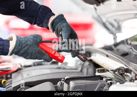 Der Mensch versucht, den Motor mit Überbrückungskabeln in Nahaufnahme zu starten. Notstart des Fahrzeugs. Hände in Winterhandschuhe halten schwarz und rot Werkzeug über offenen h Stockfoto