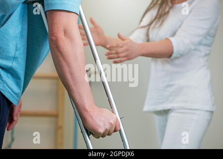 Nahaufnahme eines Mannes mit Krücke während der Rehabilitation mit einer Physiotherapeutin. Weibliche Orthopäde hilft ihrem Patienten, sich nach einer Verletzung oder einem sur zu erholen Stockfoto