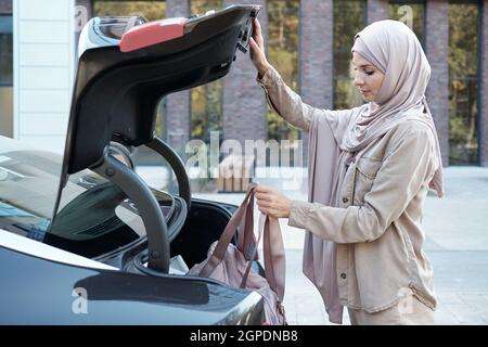 Horizontale mittlere Seitenansicht Aufnahme einer modernen muslimischen Frau, die im Freien schleift und Tasche mit Dingen in den Kofferraum des Autos legt Stockfoto
