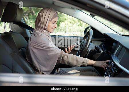 Horizontale Seitenansicht einer jungen erwachsenen muslimischen Frau, die im Auto sitzt und mit ihrem Smartphone Musik anmacht Stockfoto