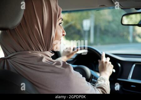 Horizontale Rückansicht – Porträtaufnahme einer modernen muslimischen Frau, die einen Hijab-Wagen trägt und in einen Seitenspiegel blickt Stockfoto