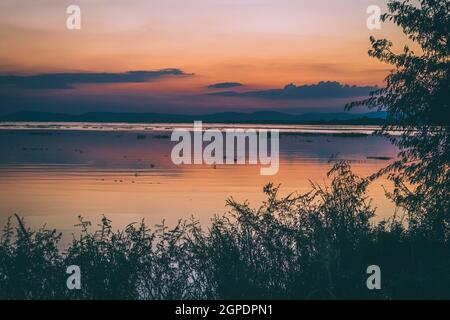 Lange Belichtung Vanille bunten Himmel und See Reflexion mit Bergen im Hintergrund. Verschwommenes wildes Gras weht im Wind im Vordergrund Stockfoto