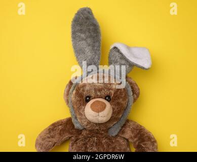 Süßer brauner Teddybär mit einer Kaninchenmaske mit langen Ohren am Kopf, lustige Feiertag-Osterkarte Stockfoto