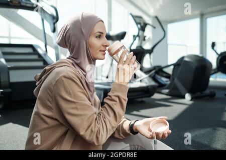 Horizontale mittlere Seitenansicht Aufnahme einer schönen muslimischen Frau, die nach dem Training im Fitnessstudio das Trinkwasser des Hijab trägt Stockfoto