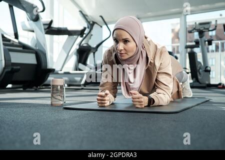 Horizontale Porträtaufnahme einer sportlichen jungen erwachsenen muslimischen Frau mit Hijab, die im Fitnessstudio Übungen zur Stärkung der Ellenbogenplanke macht Stockfoto