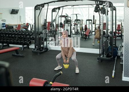 Horizontale Langaufnahme einer schönen, sportlichen muslimischen Frau, die Hijab trägt und Gewichtheben mit einer Kettlebell im Fitnessstudio macht Stockfoto