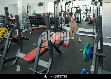 Horizontale Langaufnahme einer modernen, sportlichen muslimischen Frau, die Hijab trägt und Kniebeugen mit Kugelhantel im Fitnessstudio macht Stockfoto