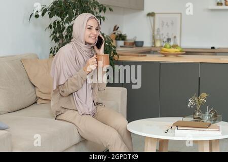 Horizontale mittellange Porträtaufnahme einer attraktiven jungen Frau mit Hijab, die zu Hause auf dem Sofa sitzt und Kaffee trinkt und am Telefon spricht Stockfoto