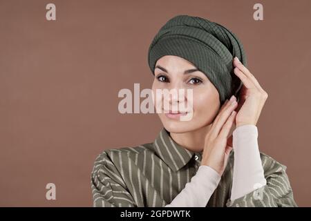 Horizontales, mittleres Nahaufnahme-Studio-Porträt einer schönen muslimischen Frau, die modisch gestreiftes Hemd trägt, das Turban auf ihrem Kopf fixiert und auf die Kamera schaut, brauner Hintergrund Stockfoto