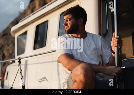 Porträt eines jungen kaukasischen bärtigen Mannes, der auf einem Deck eines Segelbootes sitzt Stockfoto