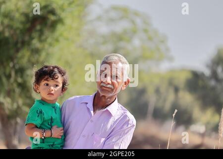 Nahaufnahme des Porträts Eines niedlichen kleinen Jungen aus einer Bauernfamilie indischer Herkunft, der im Schoß seines älteren Großvaters sitzt und die Kamera anschaut Stockfoto