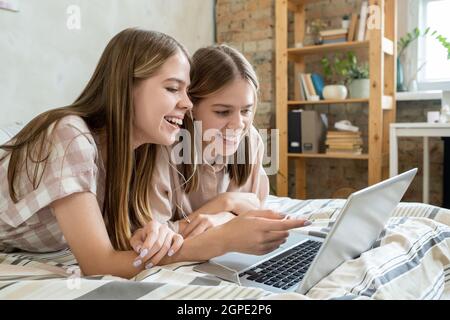 Zwei fröhliche Mädchen im Teenageralter diskutieren den Moment eines Online-Films, während sie sich auf dem Bett vor dem Laptop entspannen Stockfoto