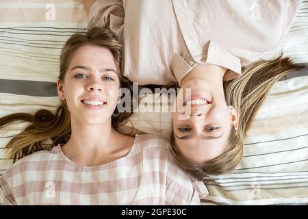 Zwei fröhliche blonde Teenager-Mädchen, die Sie betrachten, während sie sich auf dem Bett zur freien Verfügung entspannen Stockfoto