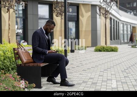 Junger bärtiger afrikanischer Geschäftsmann, der auf dem Smartphone scrollt, während er auf der Bank sitzt Stockfoto