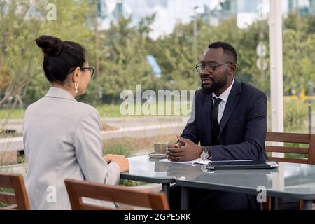 Zwei junge interkulturelle Geschäftspartner in formalwear verhandeln bei einem Treffen im Freien Stockfoto