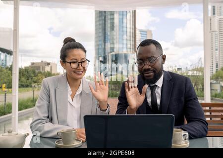 Junge, moderne Geschäftspartner begrüßen ihre Kollegen auf dem Laptop-Bildschirm mit winkenden Händen Stockfoto