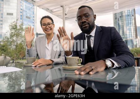 Junge erfolgreiche Geschäftspartner winken die Hände, während sie vor dem Laptop im Café im Freien sitzen Stockfoto