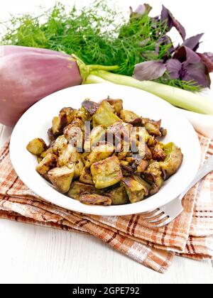 Aubergine gebraten mit Zwiebeln, Eiern und Gewürzen auf einem Teller auf Serviette, Basilikum, Lauch und Dill auf Holzbrett Hintergrund Stockfoto
