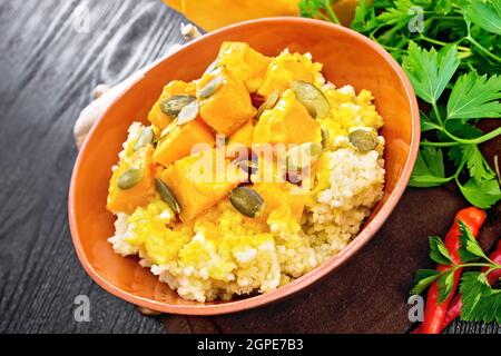 Hirsebrei mit würziger Kürbissauce und Samen in einer Tonschüssel auf Serviette, scharfer Pfeffer, Ingwerwurzel, Knoblauch und Petersilie auf dunklem Holzbrett Stockfoto